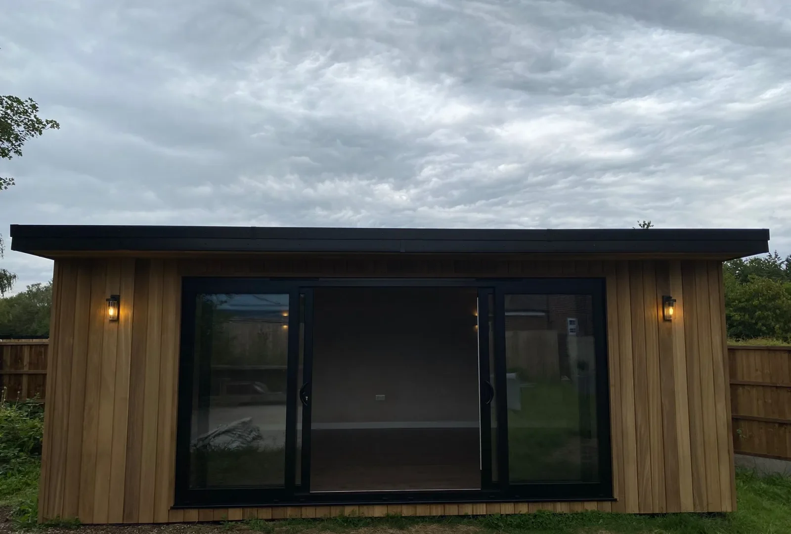Timber-clad garden room with a black flat roof trim, sliding glass doors and outdoor wall lights at dusk