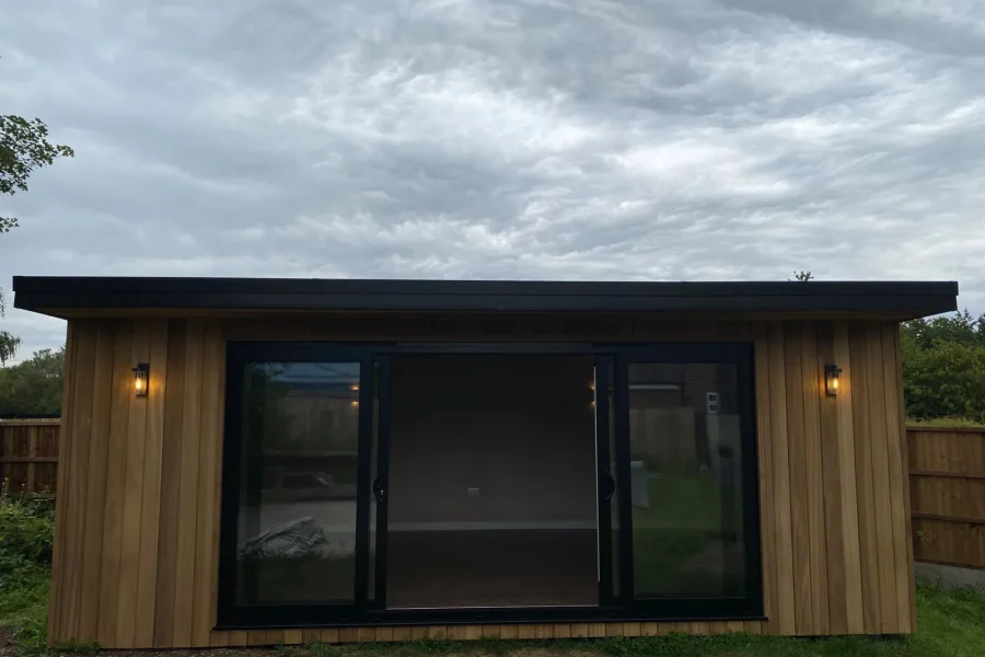 Timber-clad garden room with a black flat roof trim, sliding glass doors and outdoor wall lights at dusk