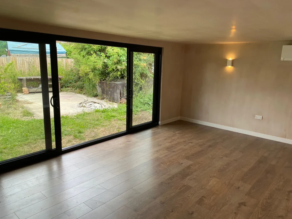 Finished garden room interior with oak-style flooring, plastered walls, a wall light and black-framed sliding doors opening onto the garden