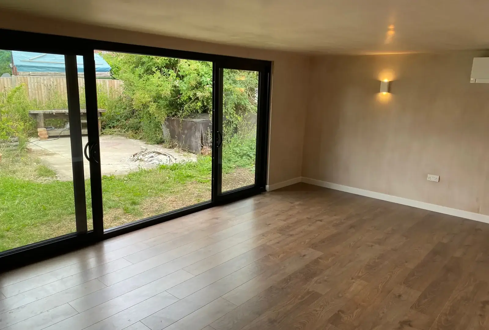 Finished garden room interior with oak-style flooring, plastered walls, a wall light and black-framed sliding doors opening onto the garden