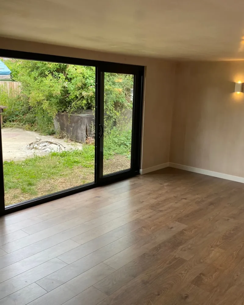 Finished garden room interior with oak-style flooring, plastered walls, a wall light and black-framed sliding doors opening onto the garden