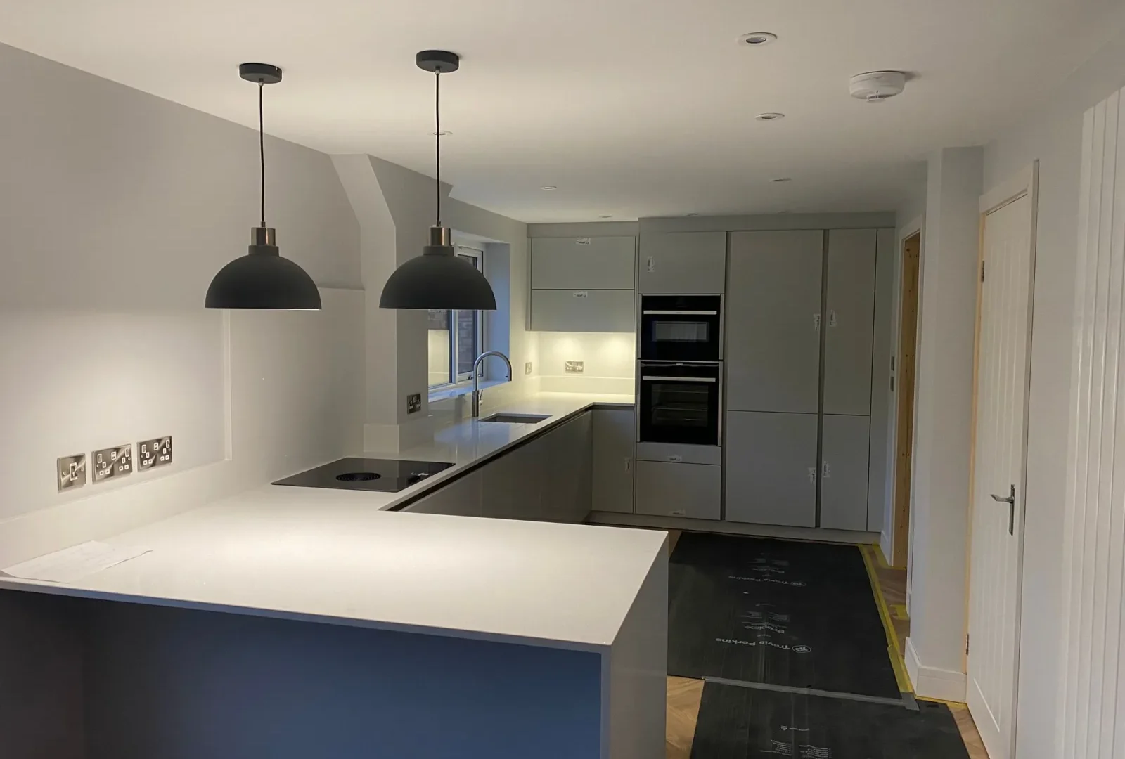 New grey handleless kitchen with white quartz worktops, a breakfast bar with pendant lights, induction hob and built-in ovens