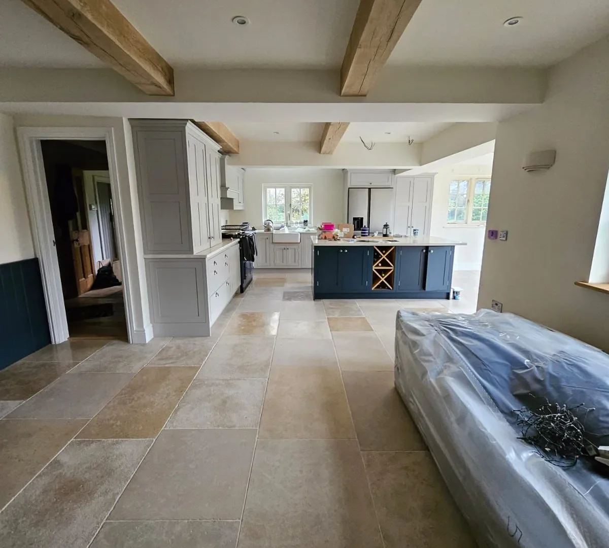 Open-plan kitchen with pale shaker units, a navy island with wine rack, oak beams and limestone flooring