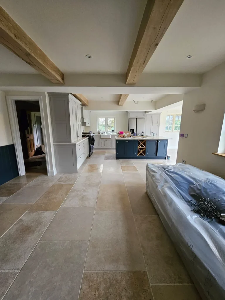 Open-plan kitchen with pale shaker units, a navy island with wine rack, oak beams and limestone flooring