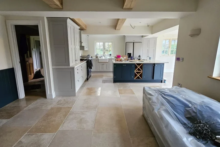 Open-plan kitchen with pale shaker units, a navy island with wine rack, oak beams and limestone flooring